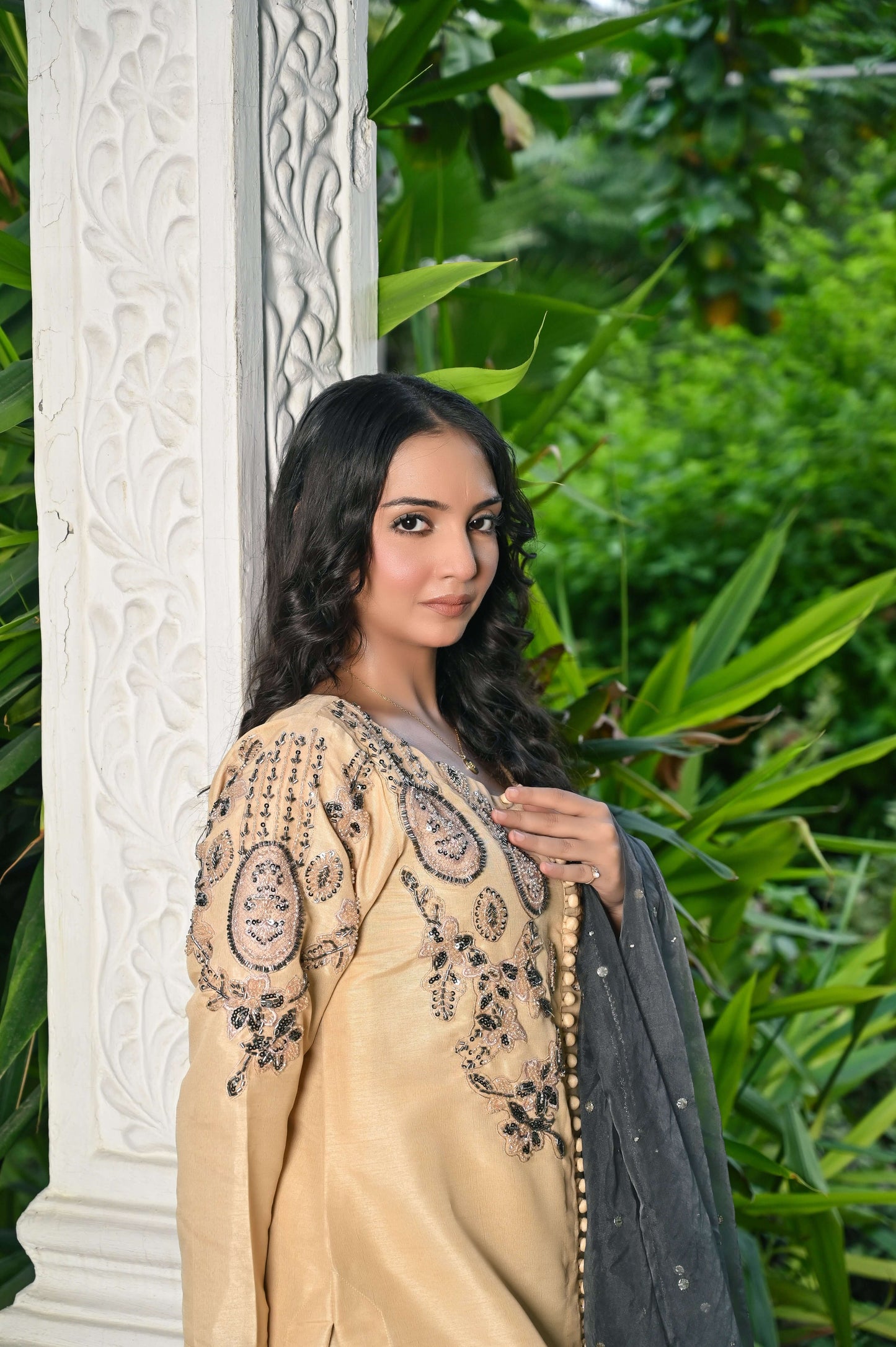 Elegant model in embroidered skin tone raw silk outfit with gray organza dupatta surrounded by lush greenery.
