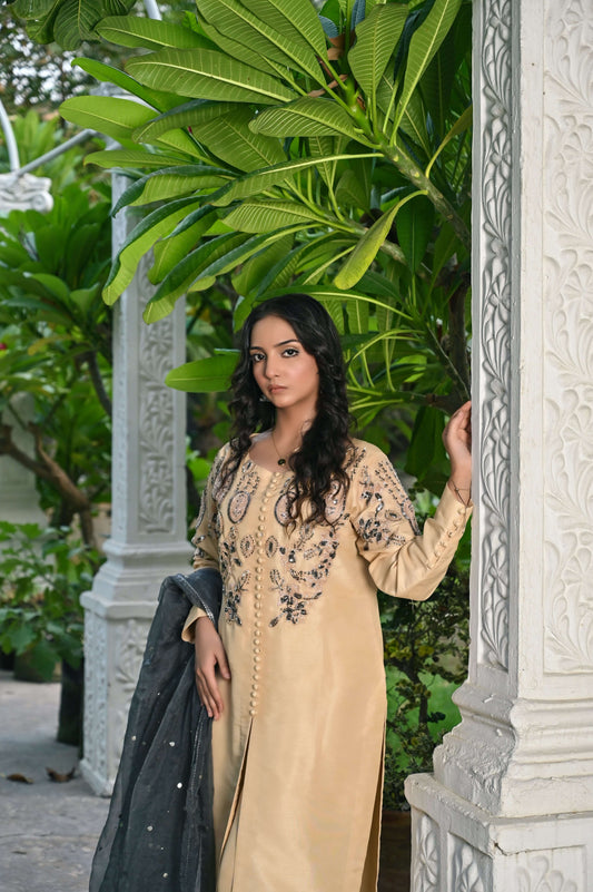 Elegant woman in a skin tone embroidered outfit with organza dupatta, surrounded by lush greenery.